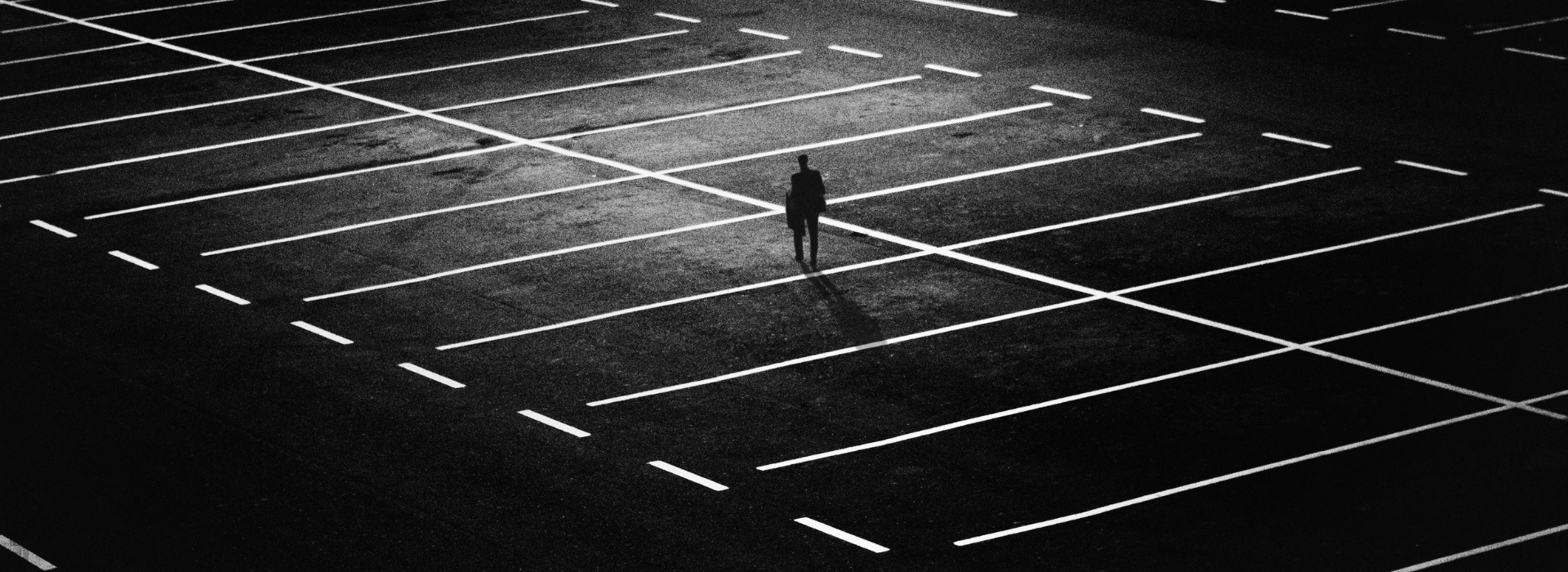 Μοναχική σιλουέτα άνδρα στέκεται στο κέντρο άδειου πάρκινγκ υπό νυχτερινό φωτισμό, συμβολίζοντας βαθιά μοναξιά και απομόνωση σε αστικό τοπίο.